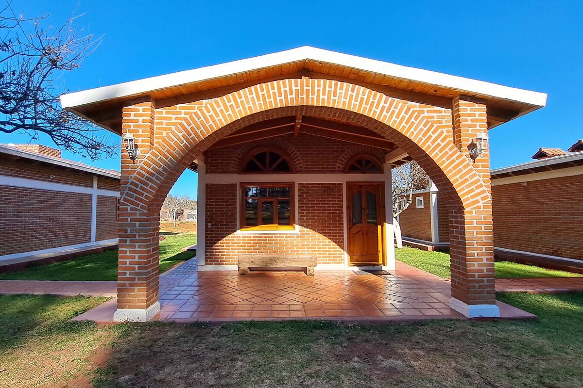 Un pabellón de ladrillo con puerta y ventanas de madera, rodeado de un césped cuidado y muros de ladrillo.