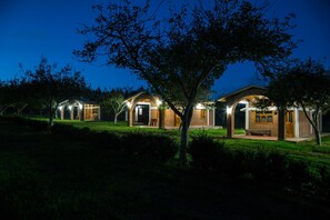 A row of modern, well-lit cabins with brick facades and arched doorways, surrounded by trees and a grassy area.