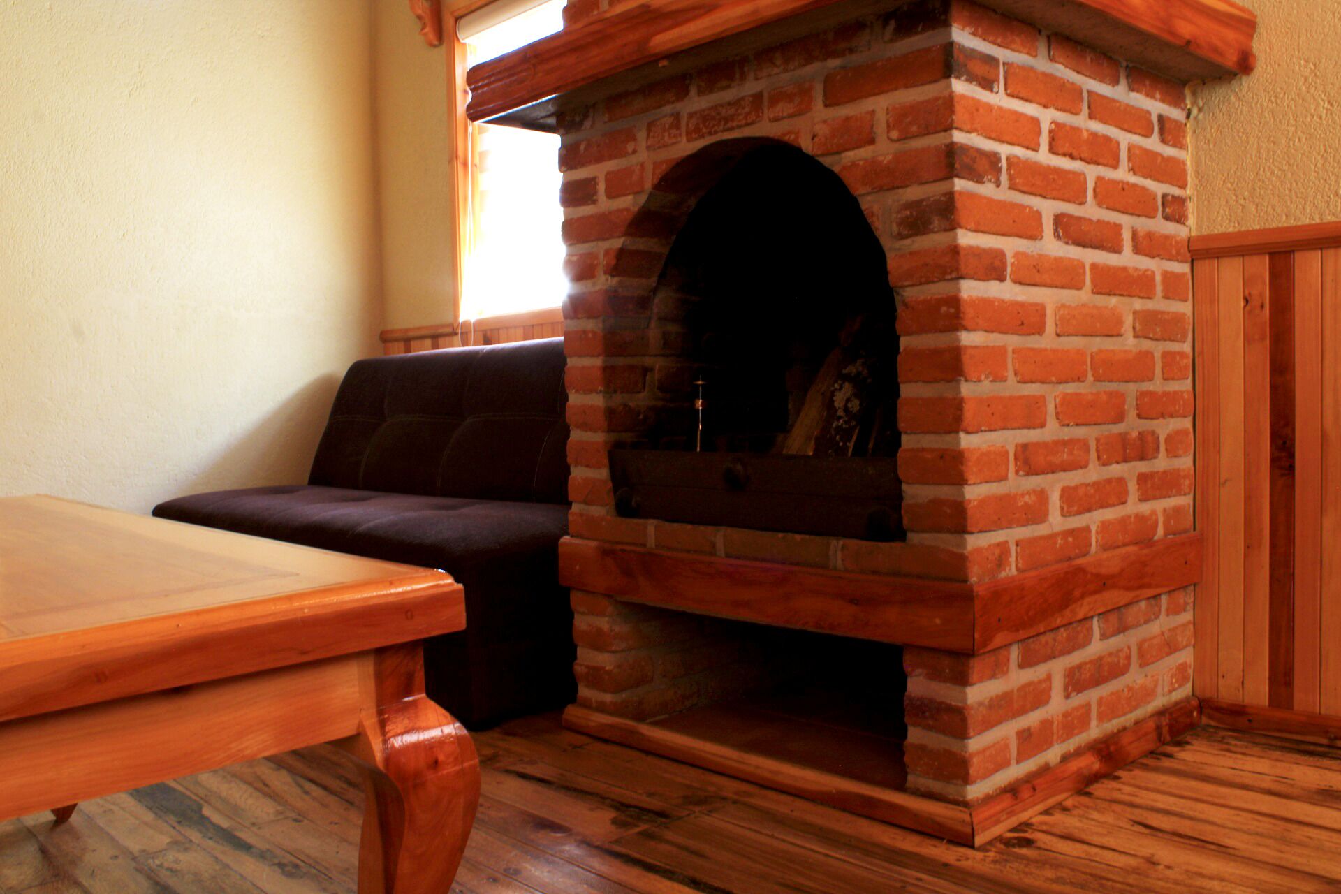 A brick fireplace with a wooden mantel, a wooden table, and a dark-colored sofa in a room with wooden flooring.