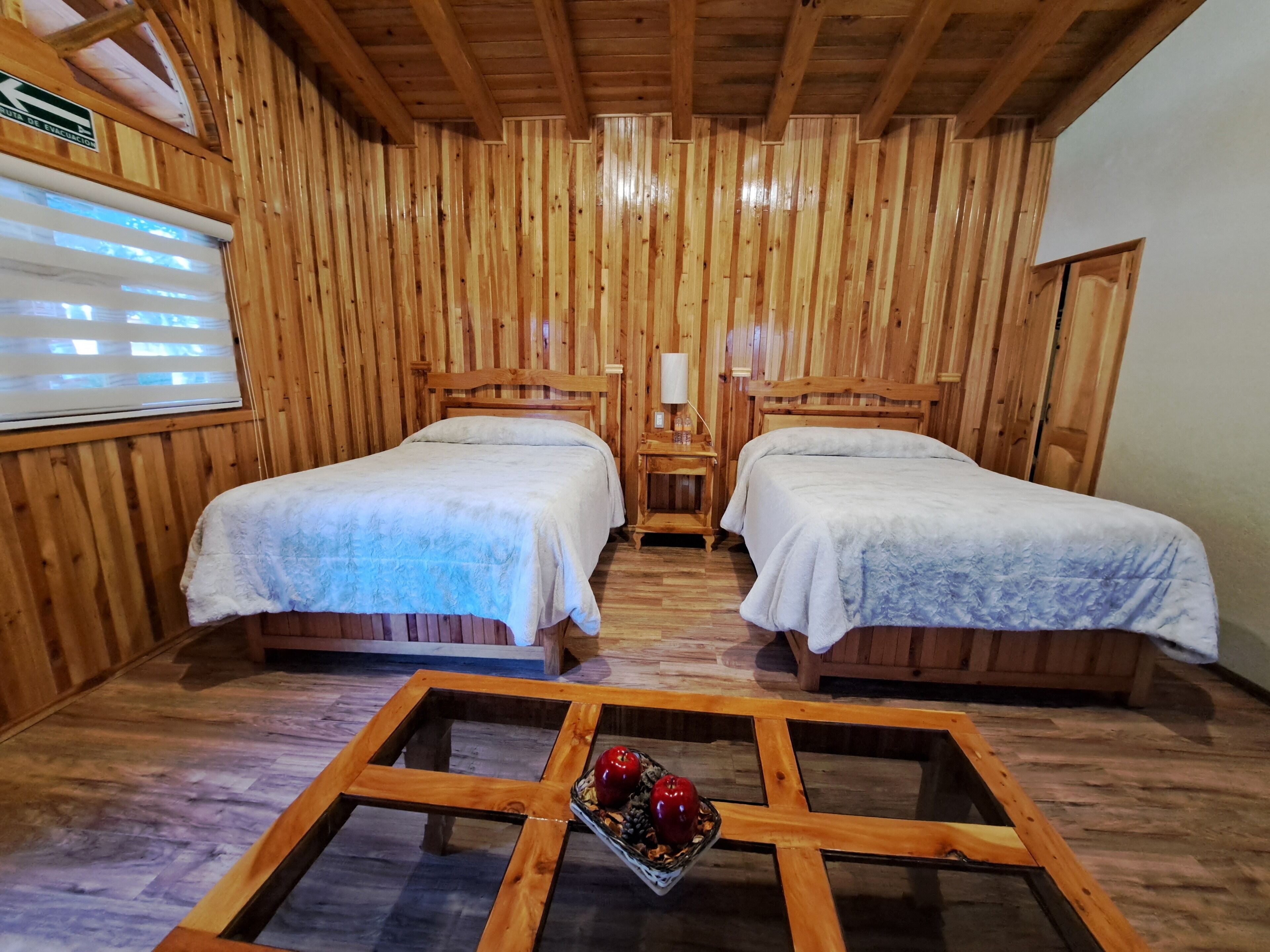 Een houten kamer met twee bedden, een kleine tafel met fruit, en een raam met jaloezieën.