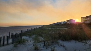 A beach view with a wooden fence, sand dunes, and buildings in the background during sunset. - Salty Snouts Retreat, Pet Friendly Home With Fire Pit in Holden Beach, NC (Holden Beach)