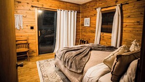 A cozy room with wooden walls, a bed with a blanket, a bench, and a door leading to a balcony. - Bear Hollow near the Buffalo River  (Jasper)