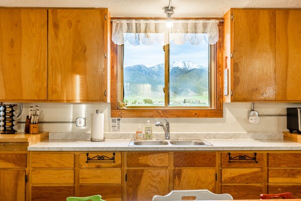 A kitchen with wooden cabinets, a sink, and a window with a view of mountains. - Solar powered farm stay near wild hotsprings! (Whitehall)