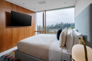 A bedroom with a large bed, a flat-screen TV mounted on the wall, and a view of a forest through a large window. - Vue Modern (Beverly Hills)