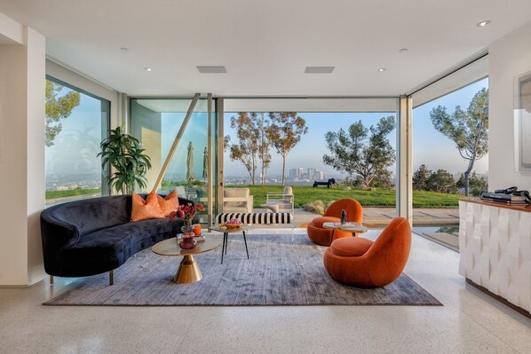 Een moderne woonkamer met een zwarte bank, oranje fauteuils, een salontafel en uitzicht op een park en stadsbeeld door grote ramen.