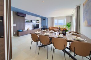 A modern dining area with a long table set for six, surrounded by six chairs. A sofa and a television are visible in the background. - Villa Wisteria with Pool (Vrbnik)