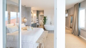 Un dormitorio moderno con una cama grande, un banco de madera, un baño con bañera y un ventanal amplio.