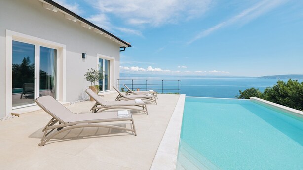 Un área de piscina moderna al aire libre con sillones de descanso, vista despejada al mar y un edificio blanco con amplias ventanas.