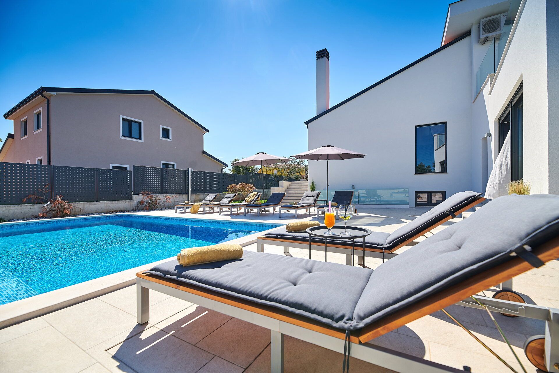A pool area with lounge chairs and umbrellas, a clear sky, and a building in the background.