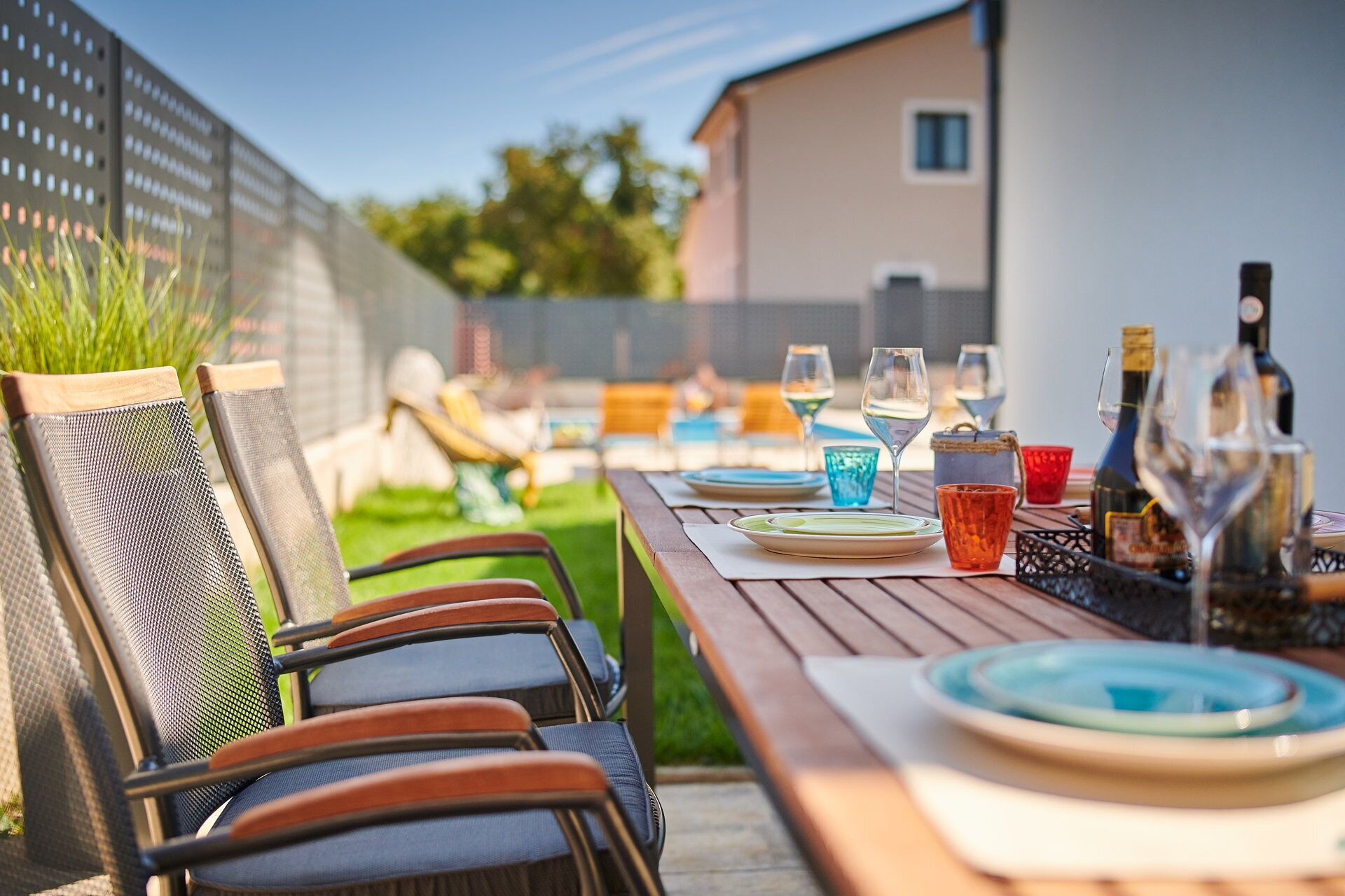Une table en bois, dressée pour un repas, avec des assiettes, des verres et des bouteilles, entourée de chaises dont les dossiers sont en tissu grillagé.