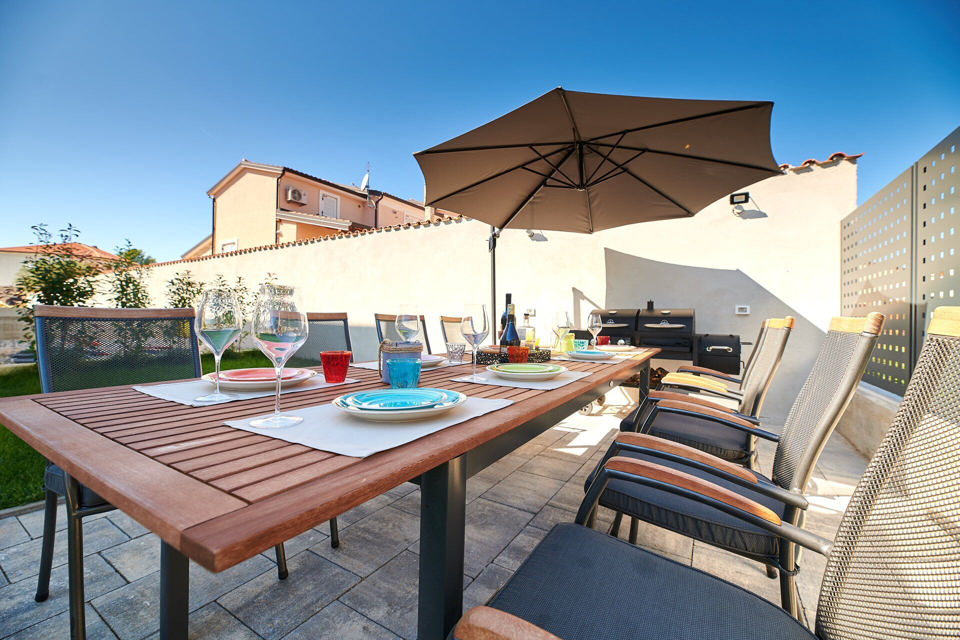 An outdoor patio with a wooden table set for dining, surrounded by chairs and an umbrella.
