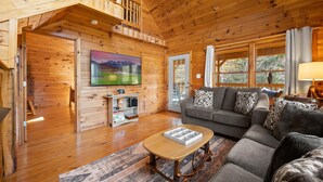 Ein Holzblockhaus-Interieur mit Wohnzimmer, einem an der Wand befestigten Fernseher und einer Treppe, die zu einer oberen Ebene führt.