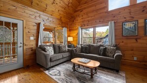 Ein gemütliches Blockhaus-Interieur mit Holzwänden, einer grauen Sofagarnitur, einem Holztisch und einem großen Fenster mit Vorhängen.