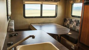 Interior compacto de una autocaravana con kitchenette, zona de comedor y ventana con vistas a un paisaje llano.