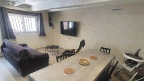 A dining area with a long table, chairs, a TV mounted on the wall, and a sofa in the background. - Pleasant house 4/6 persons free parking (Sauvian)