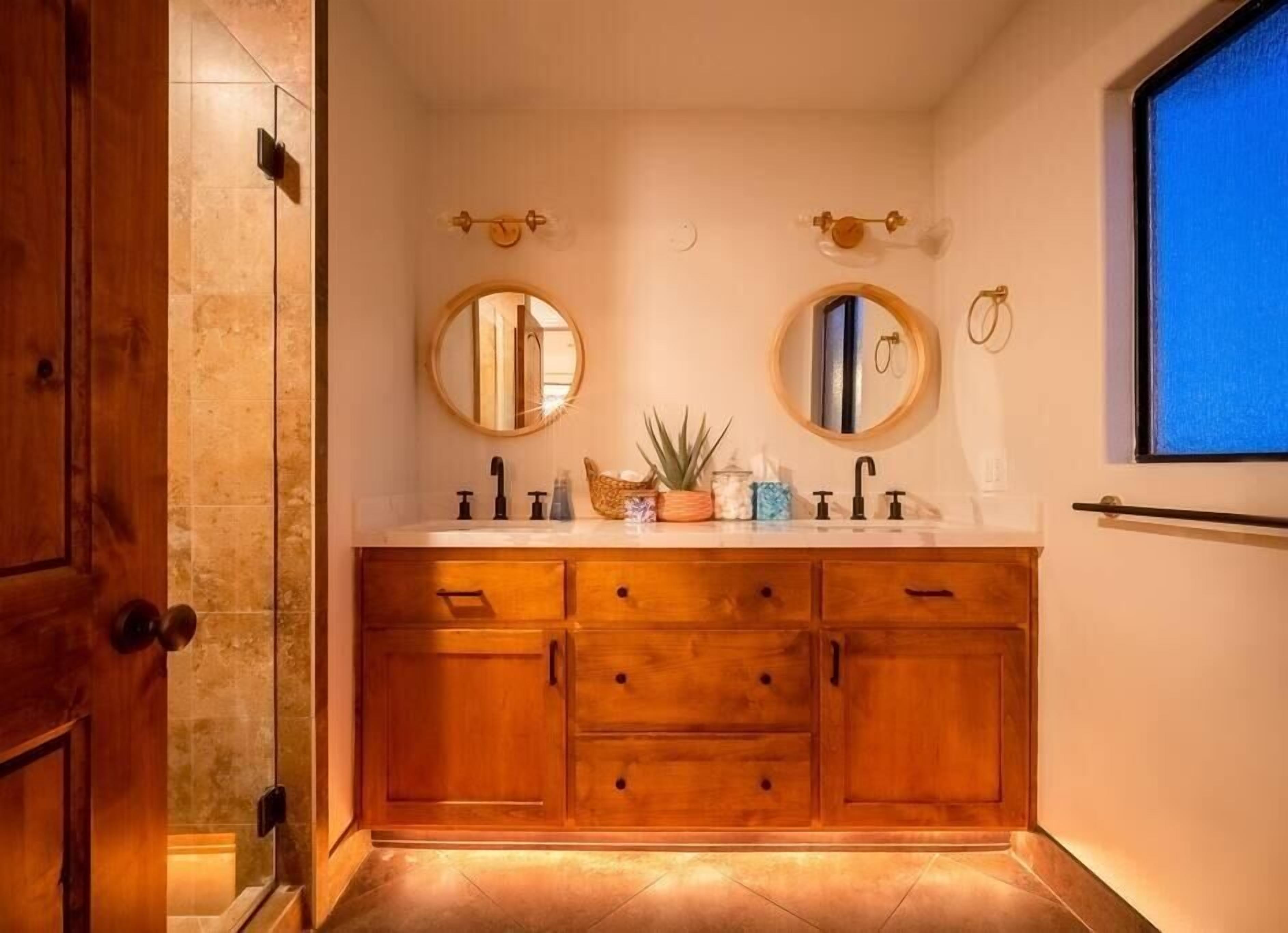 Une salle de bain avec des meubles en bois, deux miroirs ronds et une fenêtre.