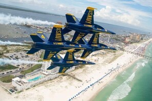 Eine Formation von US Navy Blue Angels Flugzeugen fliegt über einen Strandabschnitt, im Hintergrund sind ein Hotel und ein Steg zu sehen.