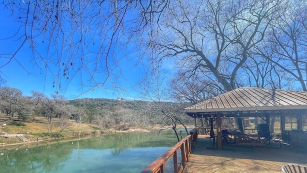 Eine Holzterrasse mit Blick auf ruhiges Wasser, mit überdachter Sitzgruppe und einem Pavillon im Hintergrund.
