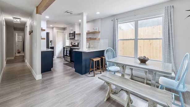 Een moderne keuken met een centraal eiland, een eethoek met een houten tafel en blauwe stoelen, en een groot raam met gordijnen.
