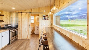 A wooden interior with a kitchen, dining area, and large windows offering a view of a grassy field and mountains.