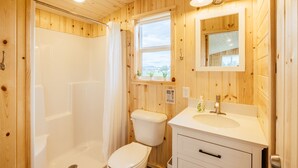 A wooden-paneled bathroom with a shower, toilet, sink, and a window offering a view.