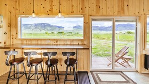 A wooden interior with a bar area, four stools, and a large window offering a mountain view.