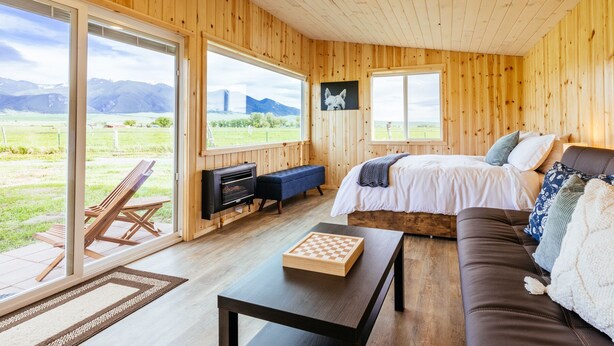 A cozy room with a bed, a sofa, a table, and a bench. There are large windows offering a view of the outdoors.