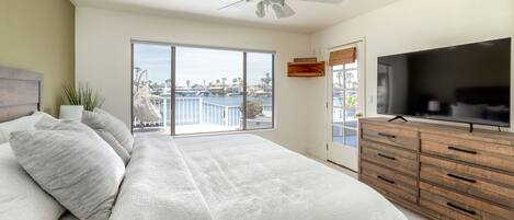 A bedroom with a bed, a ceiling fan, a TV, a wooden dresser, and a view of a marina through a sliding door.