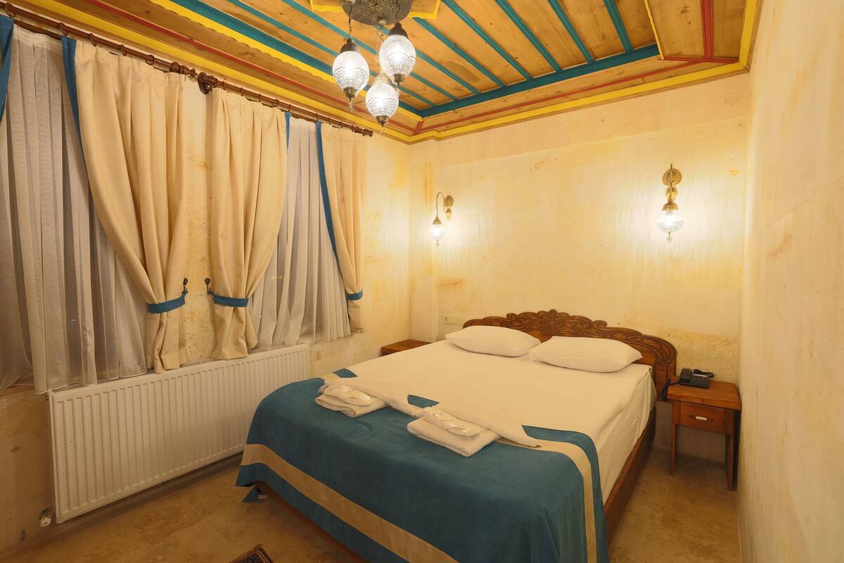 Un dormitorio con techo de madera, una cama con colcha azul y blanca, dos lámparas de pared, una mesita de noche con un teléfono y una ventana con cortinas.