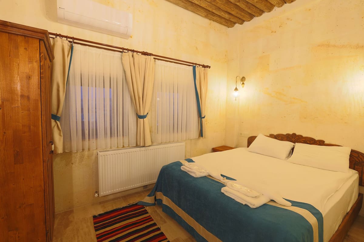 Habitación de hotel con puerta de madera, cama con ropa blanca, alfombra azul a rayas y ventana con cortinas beige.