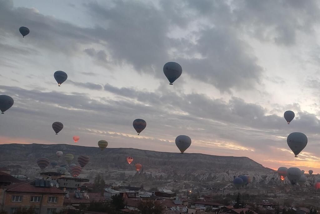 A landscape with multiple hot air balloons in the sky, a mountainous backdrop, and buildings at the base.