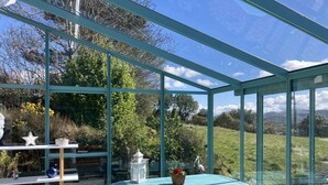 Ein Wintergarten mit Glasdach, einem blauen Tisch, weißen Stühlen und einem kleinen Gartenbereich mit Statue und sternförmiger Dekoration.