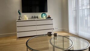 A modern living room with a glass coffee table, a white cabinet with decorative items, a flat-screen TV mounted on the wall, and a large window with sheer curtains.