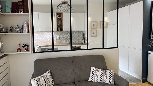 A modern living room with a grey sofa, two patterned cushions, a bookshelf with various items, and a kitchen area visible through a window.