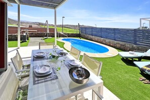 A poolside dining area with a table set for two, a small pool, and a stone wall in the background.