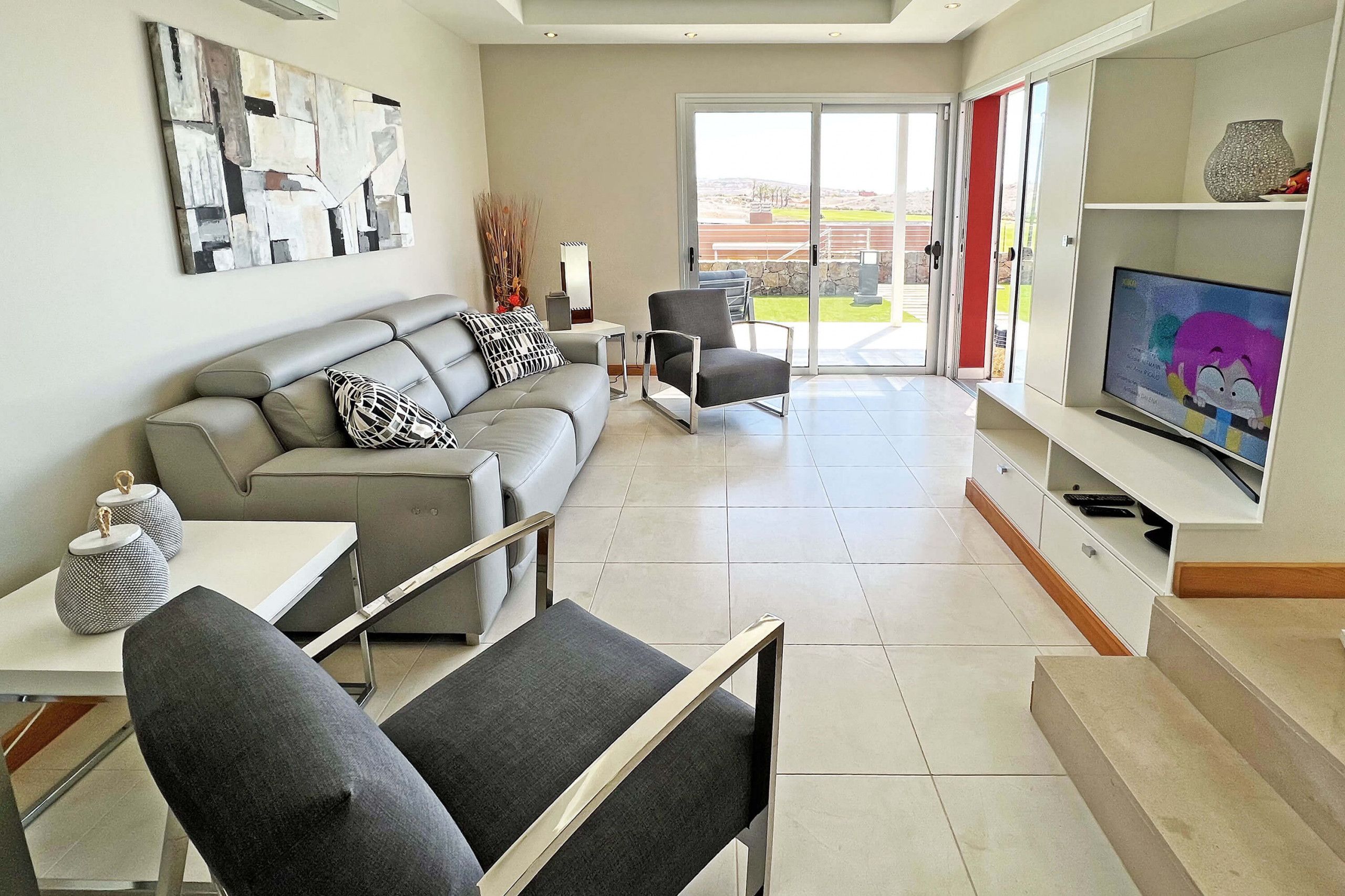 A modern living room with a grey sofa, a grey armchair, a small table with decorative items, and a television on a white stand.