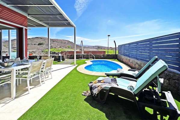 Uma área de piscina com hidromassagem, um alpendre coberto com mesa e cadeiras, e vista para uma paisagem montanhosa.