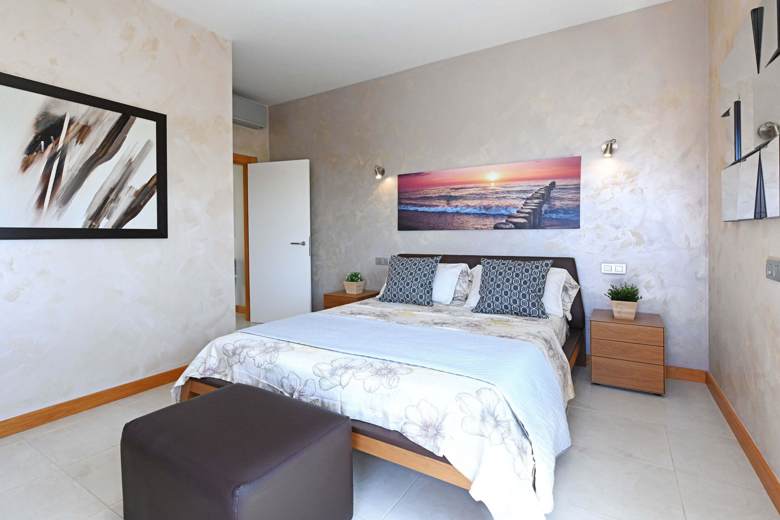 A bedroom with a bed, a nightstand, a bedside table, a chair, and a wall-mounted picture.