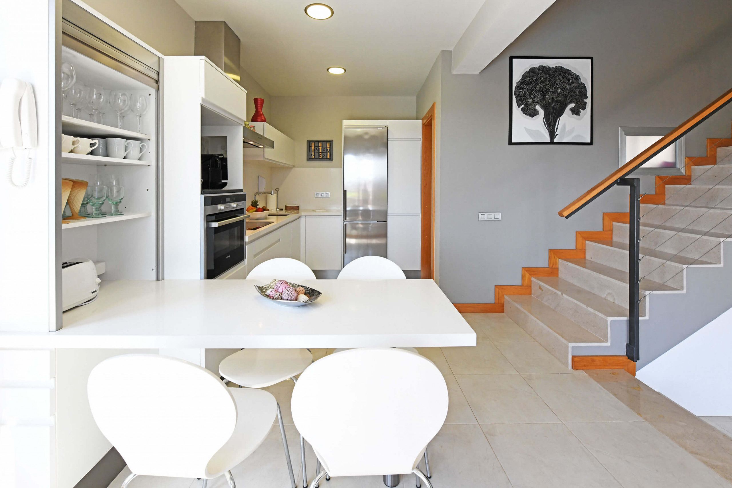 Una cocina moderna con armarios blancos, un refrigerador de acero inoxidable y un comedor con sillas blancas.