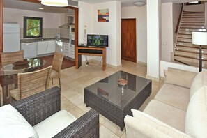 A living room with a sofa, a glass coffee table, wicker chairs, a TV, a kitchen area with cabinets, and a staircase. - Salobre Morro Golf 6 (San Bartolomé de Tirajana)