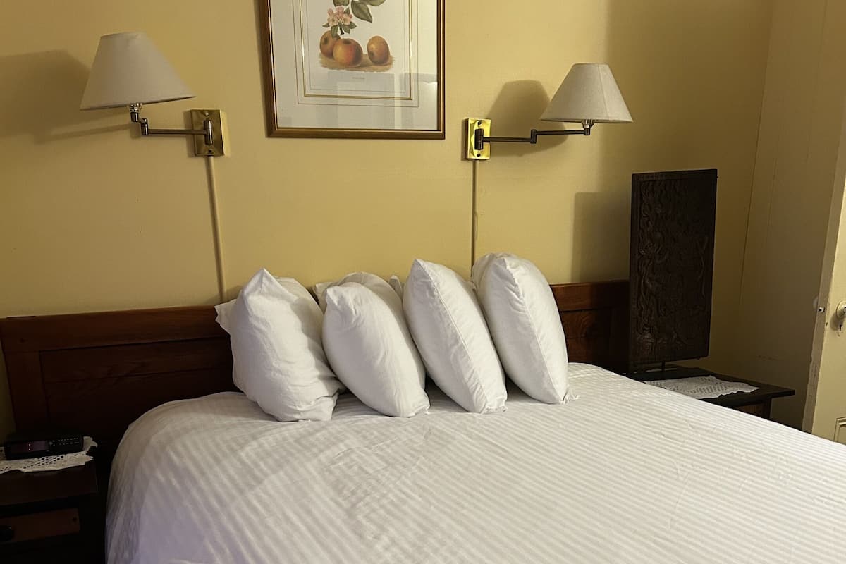 Una cama bien hecha con sábanas y almohadas blancas, un cuadro en la pared y dos lámparas fijadas a la pared.