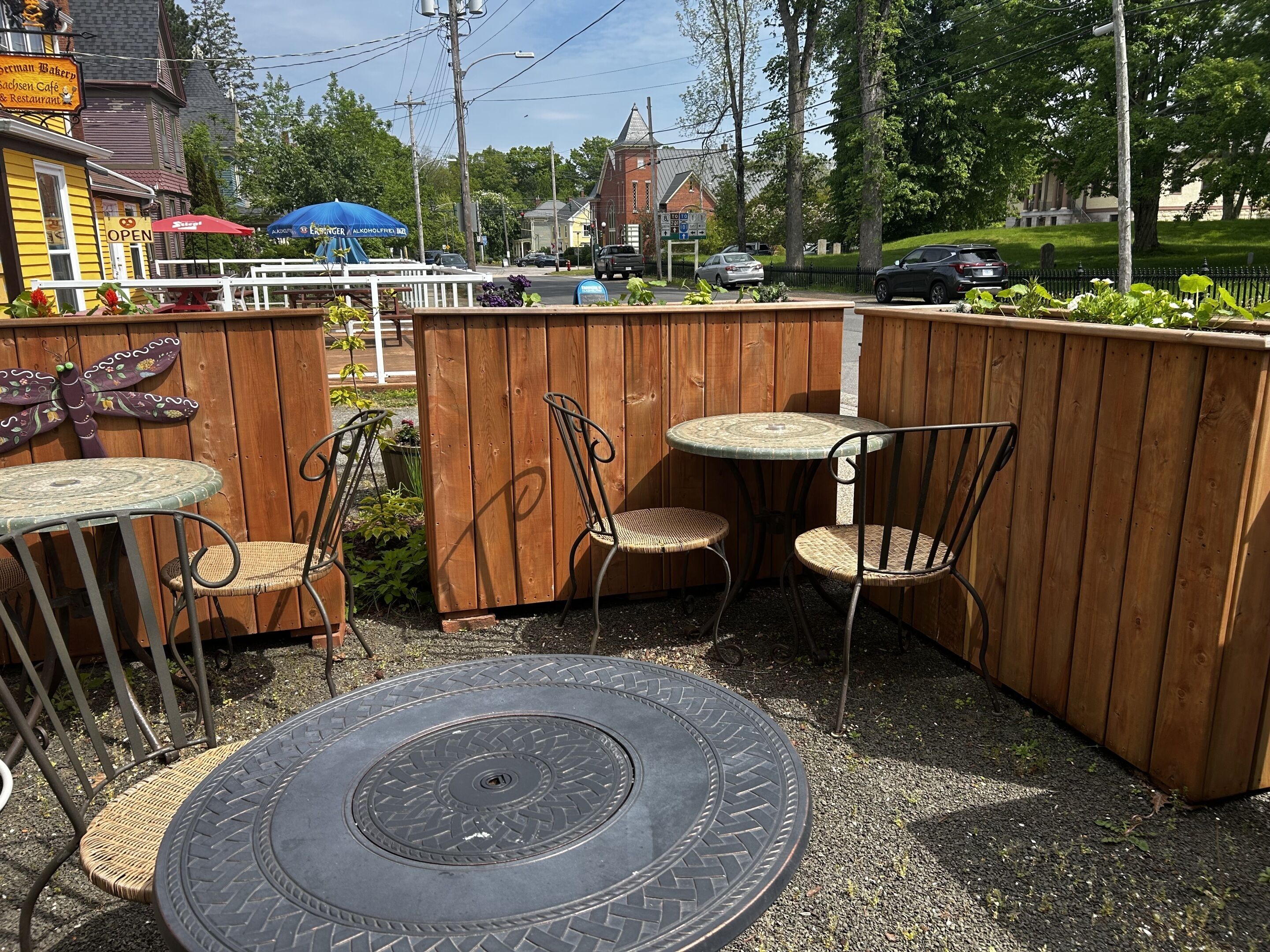 An outdoor seating area with tables and chairs, a wooden fence, and a building with a sign in the background.