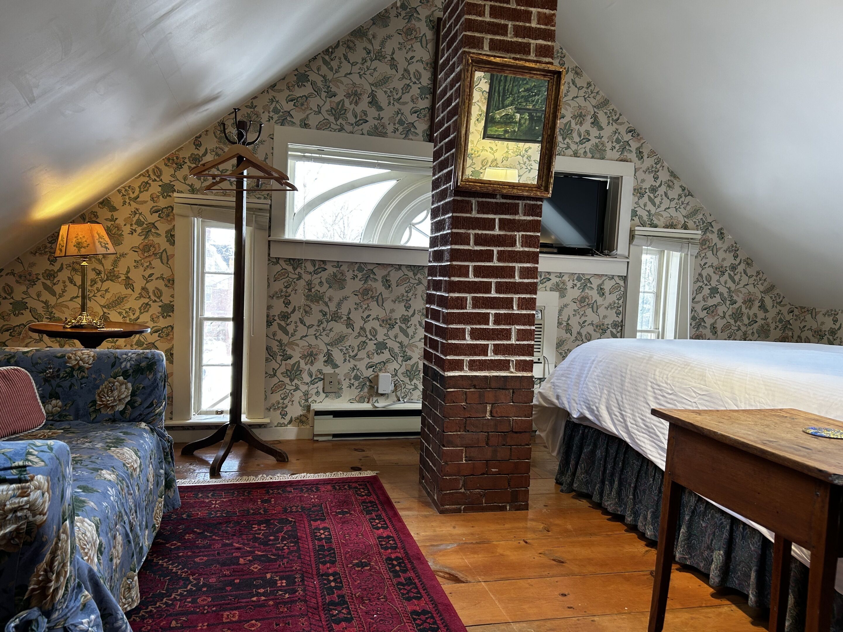 A cozy attic bedroom with a sloped ceiling, a large window, a bed, a dresser, a lamp, and a rug.