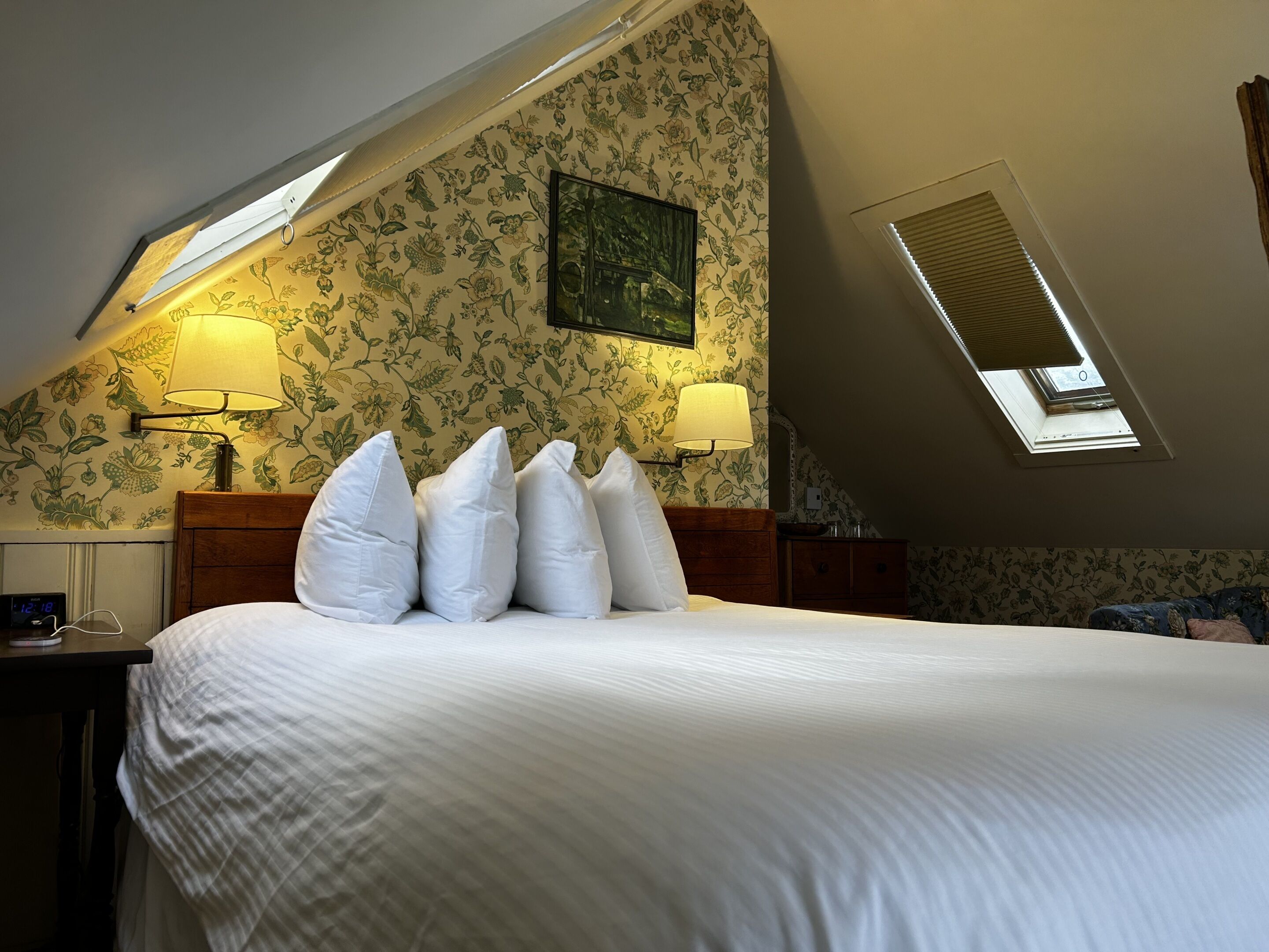 A bedroom with a large bed, two bedside lamps, floral wallpaper, a skylight, and a nightstand with a clock.