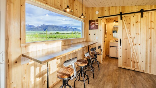 Un interior de madera con un gran ventanal que da a un paisaje montañoso, una zona de bar con taburetes y un baño a la vista.