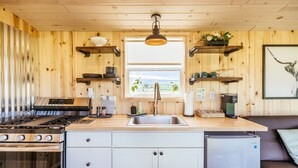 Cocina compacta con estufa, fregadero y refrigerador. Hay repisas de madera sobre la encimera que sostienen vajilla y una cafetera.