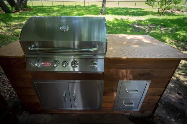 Una parrilla de acero inoxidable con cuatro quemadores y un gabinete lateral con dos cajones, sobre una plataforma de madera en un entorno exterior.