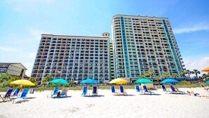 Ein Strandhotel mit mehreren Etagen, ein Sandstrand mit Liegestühlen und Sonnenschirmen sowie ein klarer, blauer Himmel.
