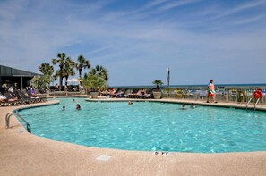 A swimming pool with lounge chairs and palm trees, surrounded by a fence and a view of the ocean. - Caribbean Resort by Elliott Bch Rentals (Myrtle Beach)