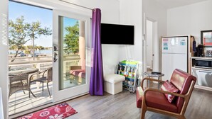 A modern room with a sliding glass door leading to a balcony, a flat-screen TV mounted on the wall, a kitchenette with a refrigerator, and a seating area with a red chair and a small table.